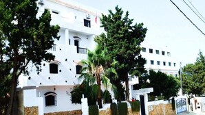 Un edificio blanco con varios balcones y una base de piedra, rodeado de árboles y un área de acceso pavimentada.