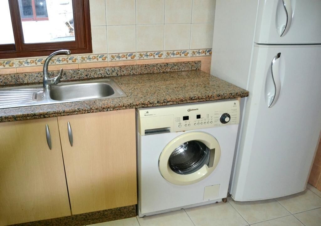 A kitchen with a granite countertop, a washing machine, a refrigerator, and a window with a view of the outside.