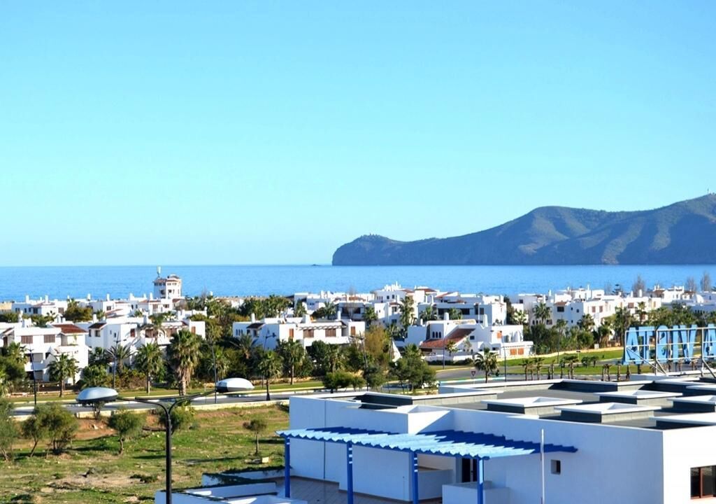 A coastal residential area with white buildings, some with blue roofs, and a clear view of the sea and a mountain in the background.