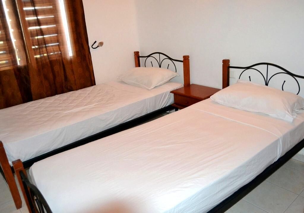 Two single beds with wooden frames and white bedding, a wooden nightstand between them, and a wooden door with blinds.