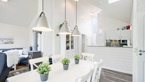 Un'area pranzo moderna e ben illuminata con un tavolo bianco, sedie grigie e lampade a sospensione. Ci sono piante in vaso sul tavolo e si intravede la cucina sullo sfondo.