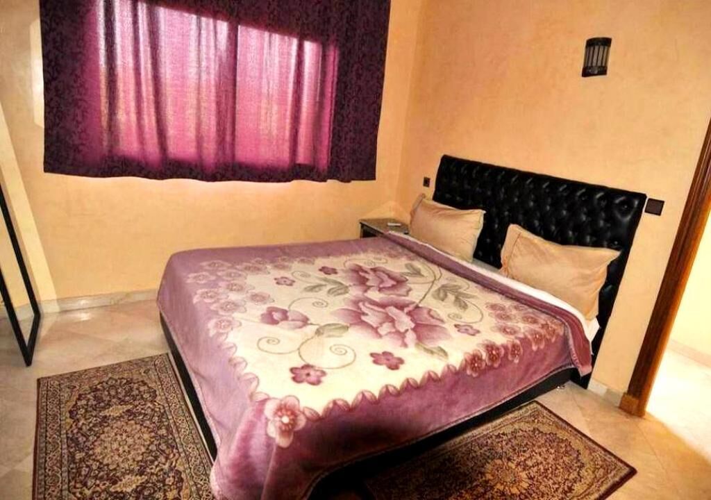 A bedroom with a bed, a nightstand, a window with curtains, and a wall-mounted light fixture.