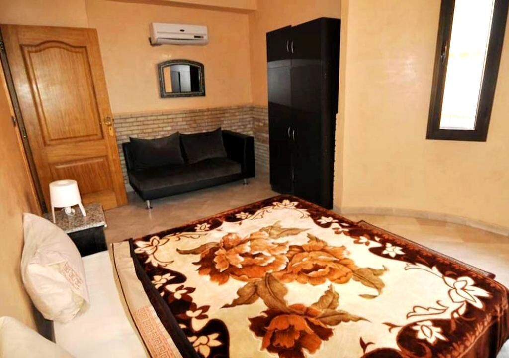 Une chambre à coucher avec un lit, un canapé de couleur sombre, une armoire noire, une petite table avec une lampe et une fenêtre avec des stores.