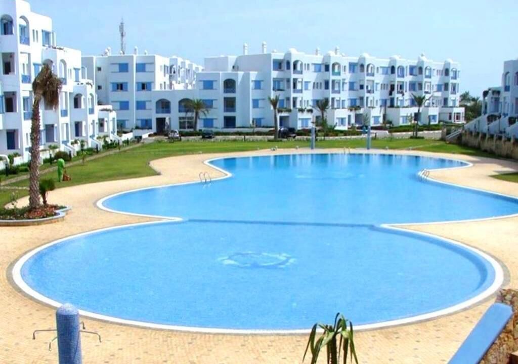 A large swimming pool with a unique shape, surrounded by a sandy area and a row of white buildings with blue accents.