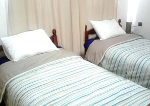 Deux lits simples avec des cadres en bois et des oreillers blancs, un rideau beige et une petite table avec une lampe.
