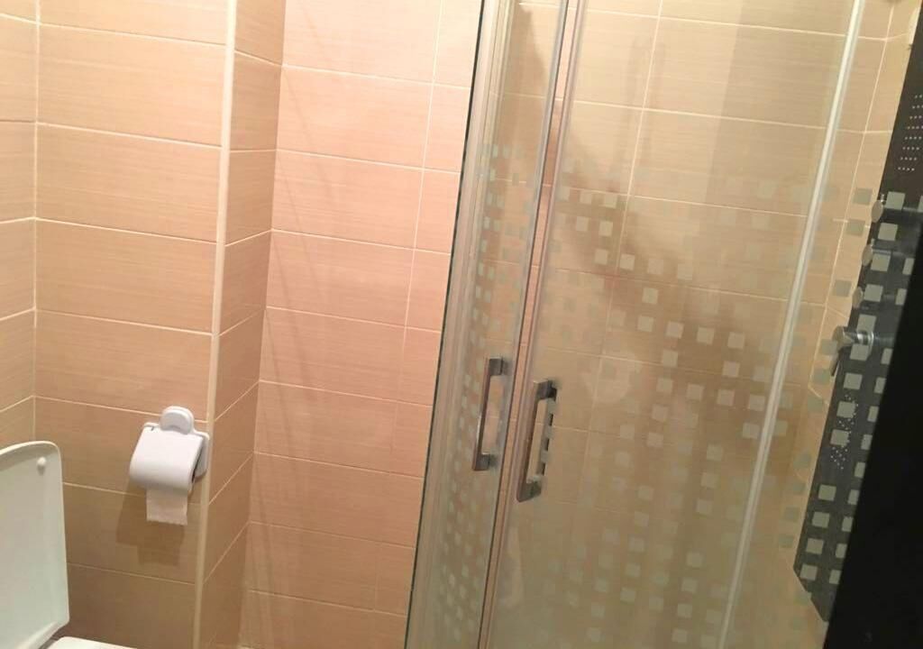A shower area with a glass door, beige tiled walls, and a toilet with a paper towel dispenser.