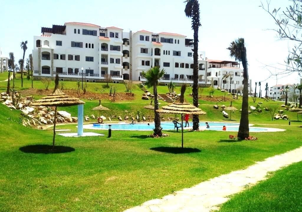 Un hotel con varios edificios blancos, piscina y zona verde con palmeras.