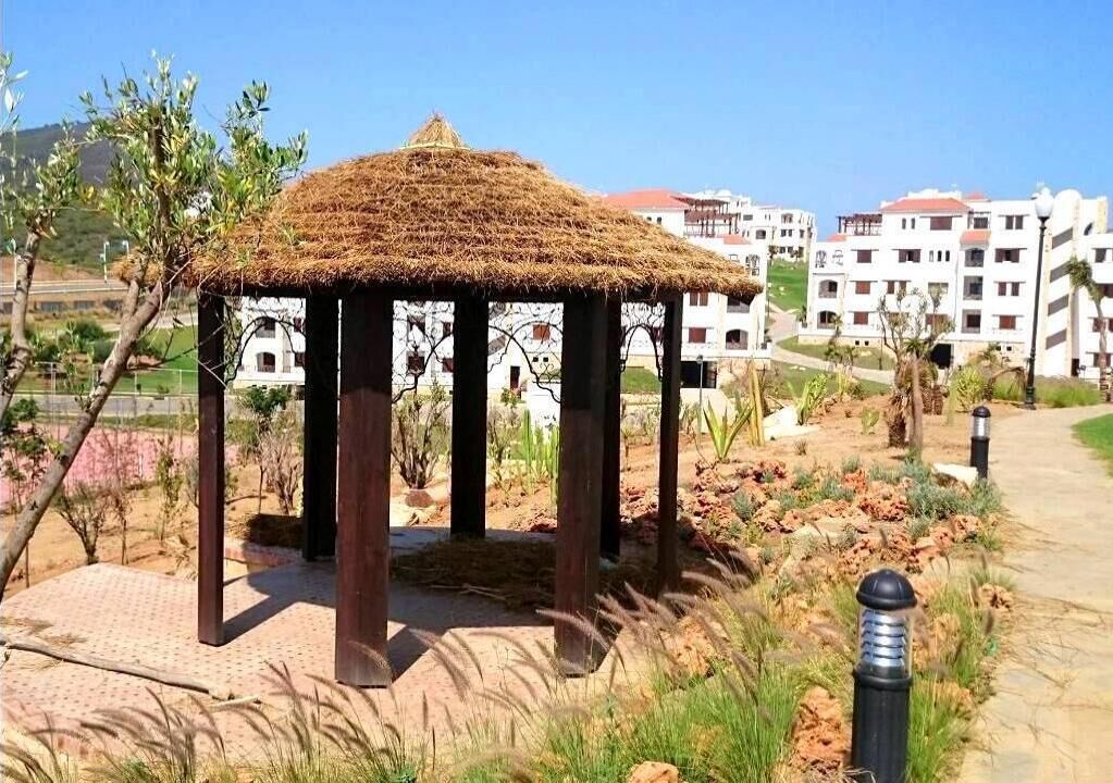 Un pabellón con techo de paja en un área ajardinada con un sendero pavimentado, rodeado de edificios y vegetación.
