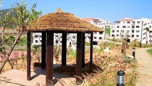 A gazebo with a thatched roof in a landscaped area with a paved walkway, surrounded by buildings and greenery. - Lilac's Garden Residence 2194 (Cabo Negro)