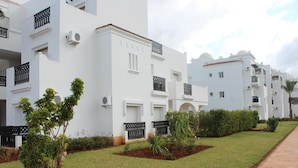 Fileira de edifícios brancos com varandas, um passeio pavimentado e pequenas áreas ajardinadas com plantas.