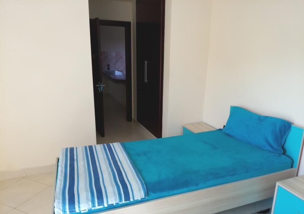 Habitación individual con una cama, un edredón a rayas, una almohada azul y una mesita de noche de madera.