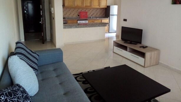A living room with a sofa, coffee table, and TV stand. A kitchen is visible in the background with wooden cabinets and a refrigerator. - Atlantic Ocean Residence 3041 (Asilah)