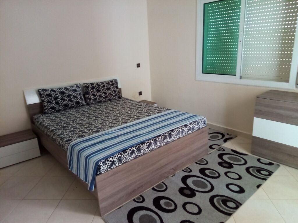 A bedroom with a bed, a window with blinds, a bedside table, and a patterned rug.