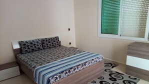 A bedroom with a bed, a window with blinds, a bedside table, and a patterned rug. - Atlantic Ocean Residence 3041 (Asilah)