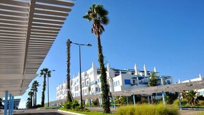Een hotel aan de kust met een modern design, voorzien van een overdekte doorgang, palmbomen en een helderblauwe lucht.