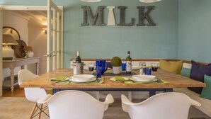 A dining area with a wooden table set for two, white chairs, and a wall-mounted sign that reads "MILK". - The Old Dairy, Great Bircham, Norfolk (Bircham)