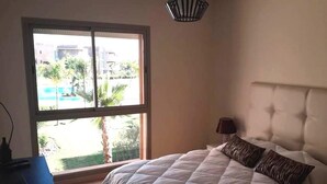 A bedroom with a large bed, a nightstand, a lamp, and a window with a view of a pool and greenery. - Residence Marrakech Prestigia Opal 2999 (Marrakech)
