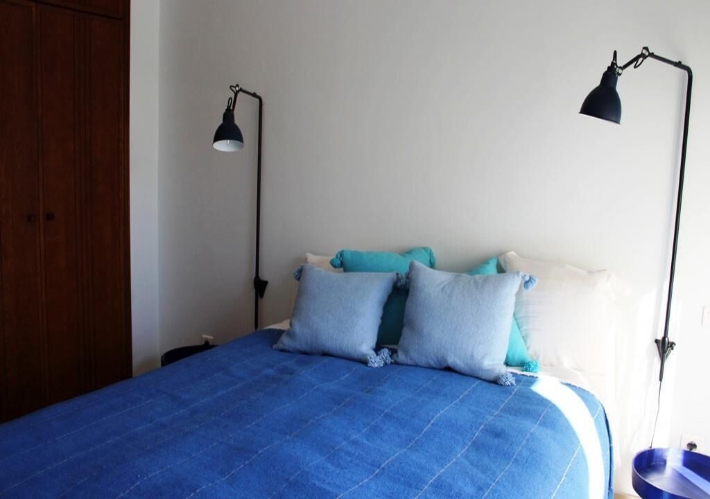 A bedroom with a blue bedspread, two pillows, a wooden wardrobe, and two wall-mounted lamps.