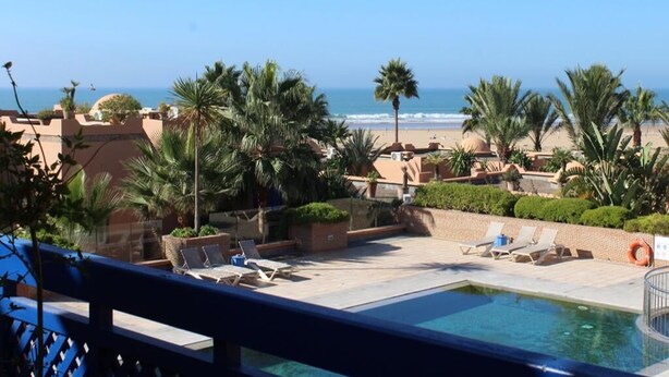 Ein Poolbereich mit Liegestühlen und Palmen, mit Blick auf einen Strand.