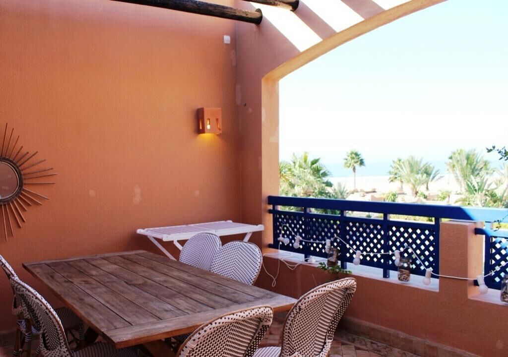Une terrasse extérieure avec une table en bois et des chaises en osier, un élément décoratif mural et une vue sur la plage.