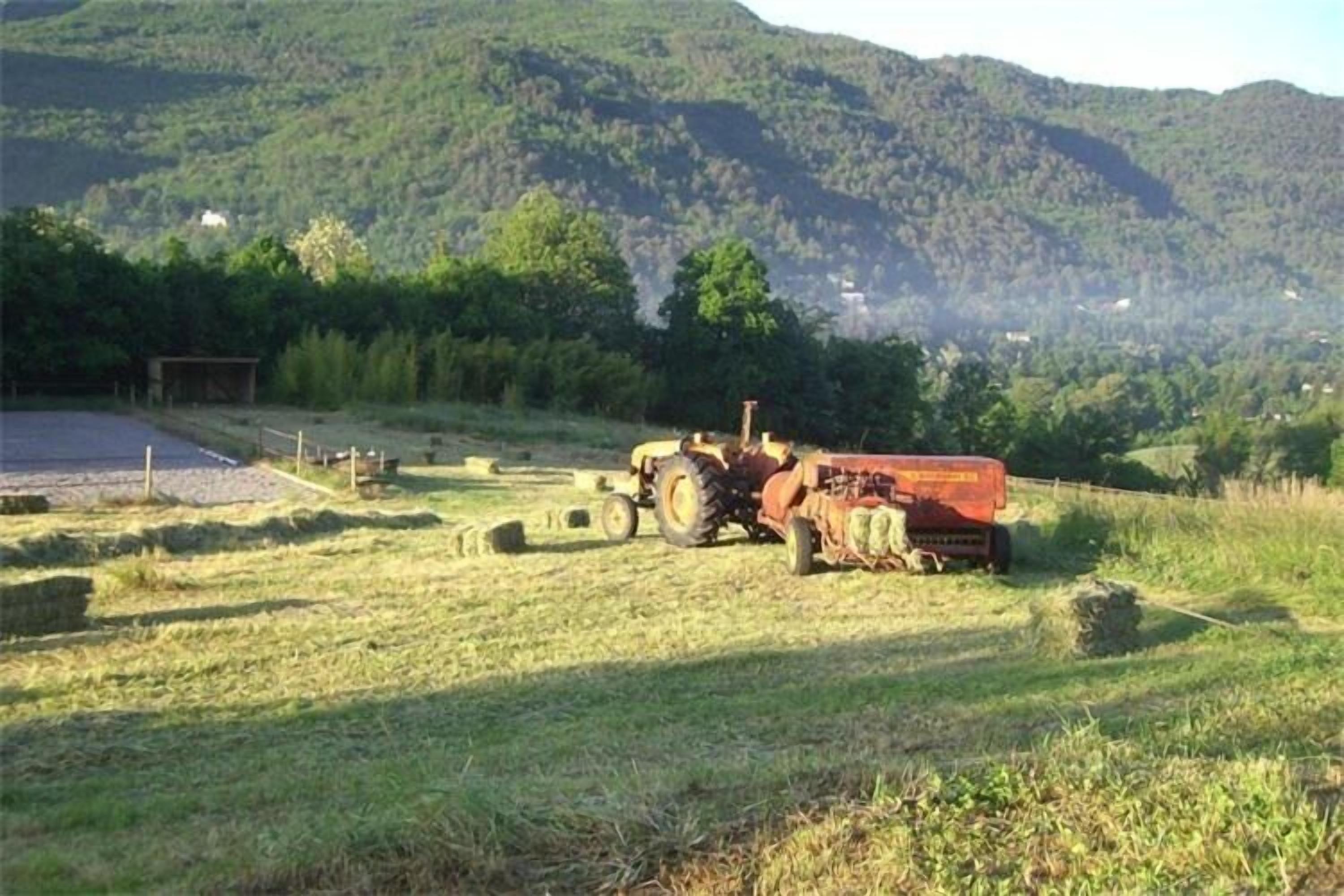 A tractor in a field with hay bales, a shed, and a mountainous backdrop.