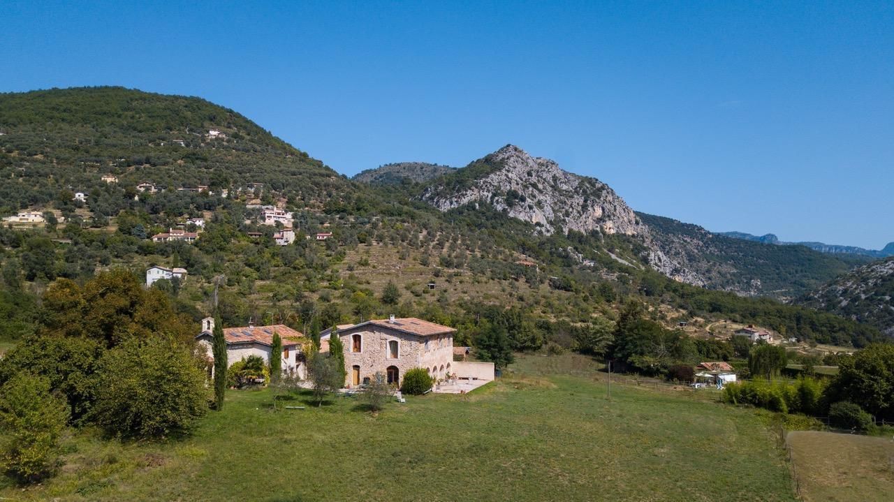 Un paisaje rural con dos construcciones de piedra, una con techo rojo y otra con techo gris, rodeado de vegetación y colinas.
