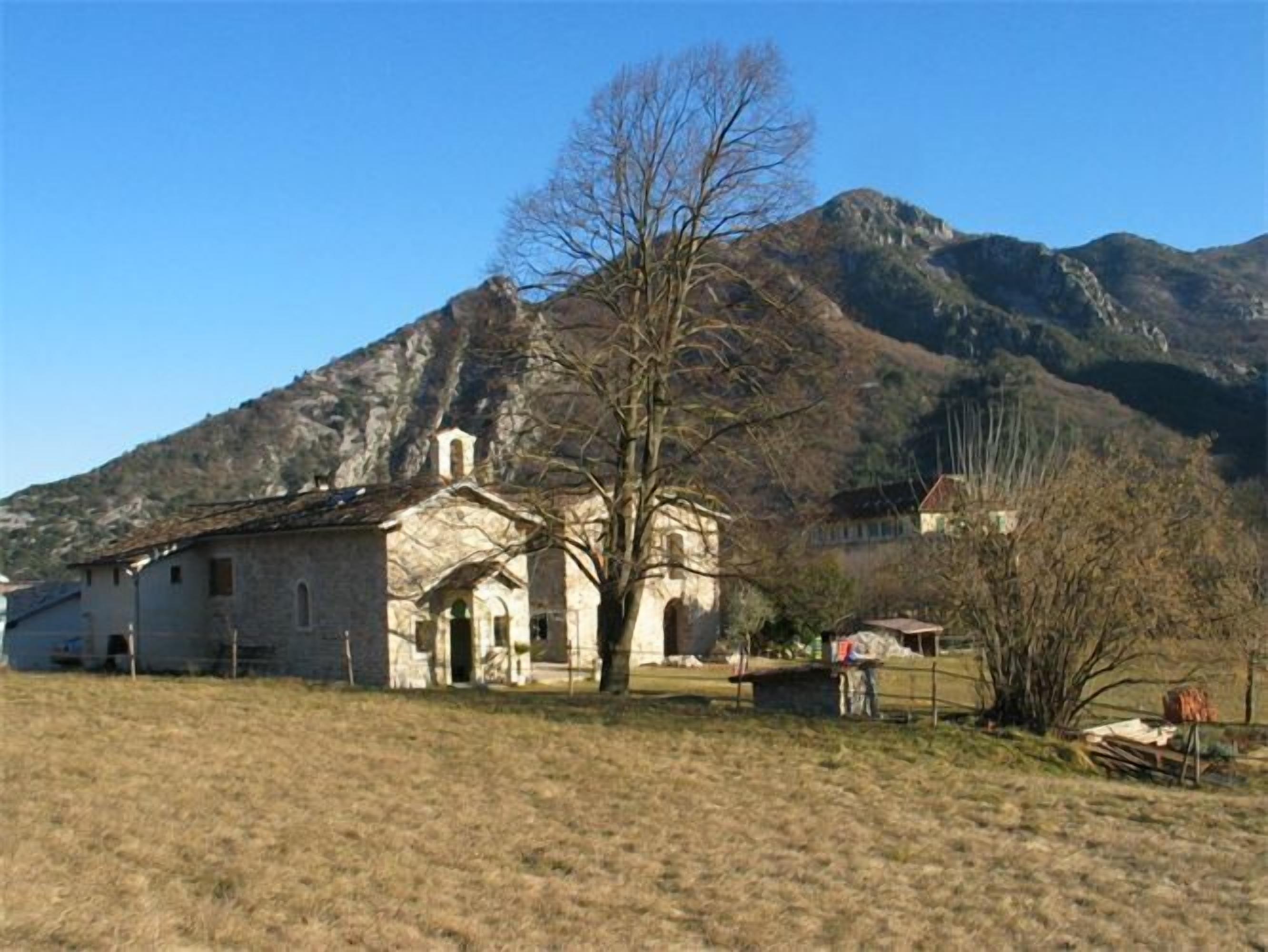 A stone building with a bell tower, surrounded by a grassy field and mountains in the background.