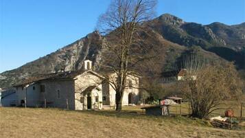 A stone building with a bell tower, surrounded by a grassy field and mountains in the background.