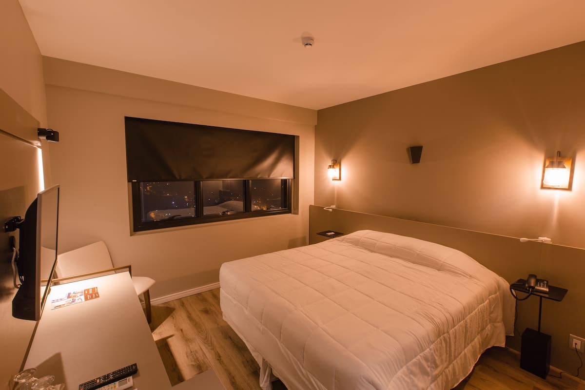 A hotel room with a large bed, a desk with a chair, a TV, and a window with a view of the city at night.