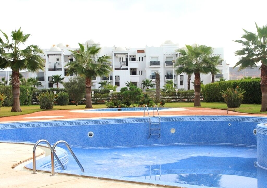 A swimming pool with a blue mosaic border, surrounded by palm trees and a building with white walls and balconies.