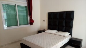 Un dormitorio con una cama, dos mesitas de noche, un ventanal con persianas y una cortina roja.