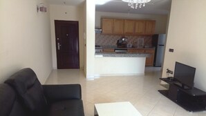 Una sala de estar con un sofá negro, una mesa de centro blanca y un mueble para televisor. Se ve una cocina al fondo con armarios de madera y piso de baldosas.