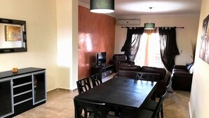 A dining area with a long table and chairs, a television, and a sofa. - The Greens Residence 3613 (SAIDIA)