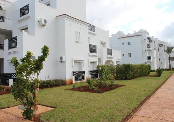 A row of white buildings with balconies, a paved walkway, and small landscaped areas with plants. - The Greens Residence 3652 (SAIDIA)