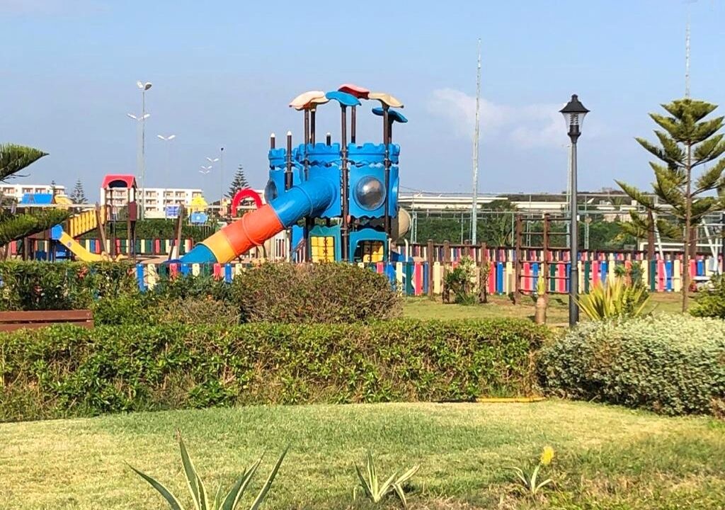 A playground with a blue and red slide, surrounded by greenery and a fence.
