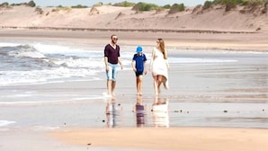Eine Familie spaziert über einen sandigen Strand, im Hintergrund ein ruhiges Meer und Dünen.