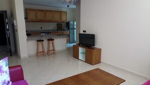 A living room with a wooden table, two purple sofas, a television on a cabinet, and a kitchen in the background. - Atlantic Ocean Residence 3043 (Asilah)