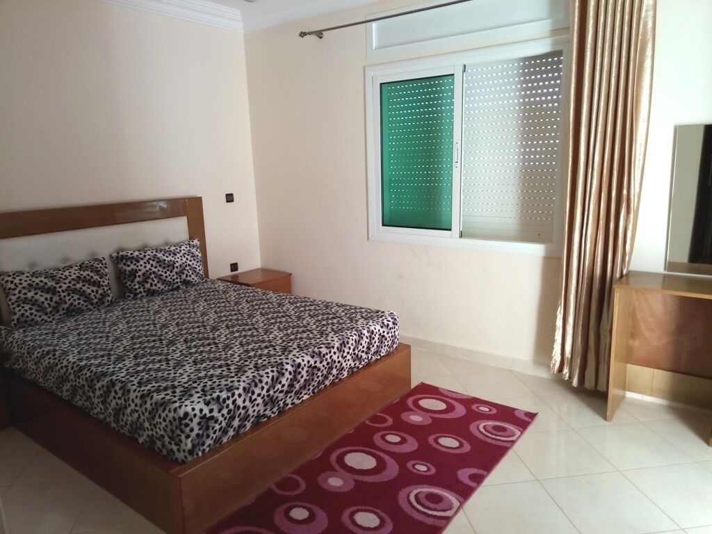 A bedroom with a bed, a window with blinds, a bedside table, and a red patterned rug.