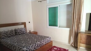 A bedroom with a bed, a window with blinds, a bedside table, and a red patterned rug. - Atlantic Ocean Residence 3043 (Asilah)