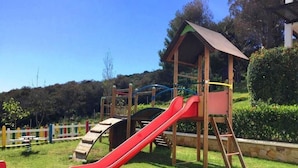 Um parquinho com um escorrega vermelho, estruturas de escalada e um abrigo de madeira.