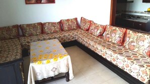 Uma sala de estar com um sofá, uma mesa com toalha de mesa florida e um quadro na parede.