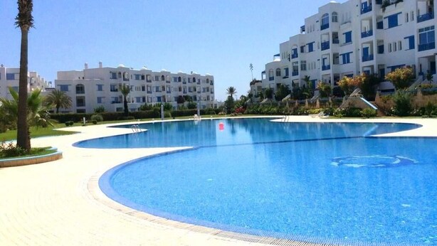 Una grande piscina trasparente circondata da un marciapiede lastricato, con edifici bianchi e palme sullo sfondo.
