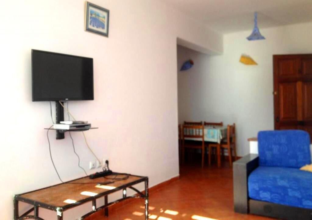 Un saló amb un televisor de pantalla plana penjat a la paret, una taula de centre de fusta, un sofà blau i una zona de menjador al fons.