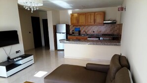 A modern living room with a brown leather sofa, a flat-screen TV, and a wooden coffee table. A kitchen with wooden cabinets and a granite countertop is visible in the background. - Atlantic Ocean Residence 3033 (Asilah)