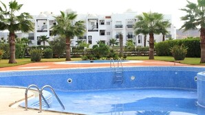 Una piscina con borde de mosaicos azules, rodeada de palmeras y un edificio con paredes blancas y balcones.