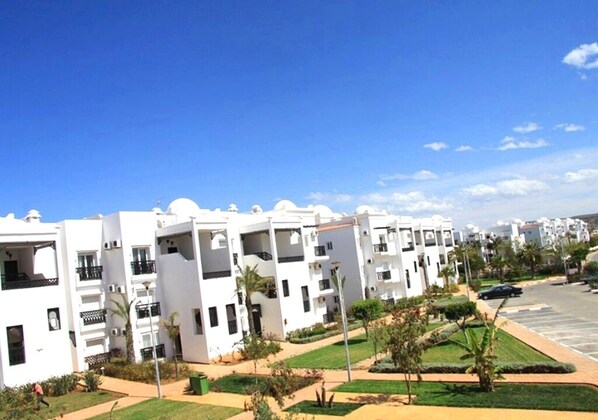 A row of white multi-story buildings with balconies, a parking lot, and a clear blue sky.