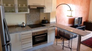 A compact kitchen with a built-in oven, a microwave, and a stovetop. There is a small dining area with a bar stool and a television. - The Greens Residence 3625 (SAIDIA)