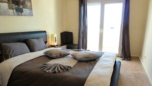 A bedroom with a large bed, a nightstand, a lamp, and a view of the outdoors through a sliding door. - The Greens Residence 3625 (SAIDIA)