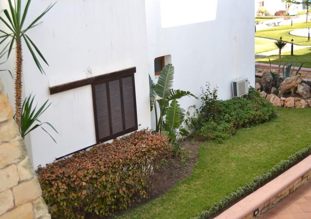 A white building with a window and a small door, surrounded by a well-maintained garden with grass, shrubs, and a palm tree.