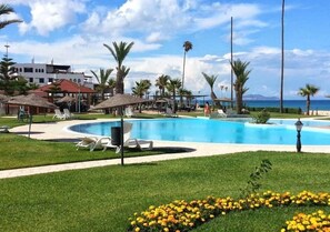 A pool area with lounge chairs, palm trees, and a building in the background. - Residence Marina Beach Mdiq 3311 (Mdiq)